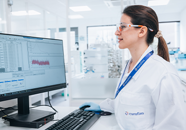 A laboratory scientist wearing a white lab coat and blue gloves is seated at a workstation at Eurofins BioPharma Product Testing laboratory, analyzing data on a computer screen. The display shows scientific graphs and tables, suggesting the evaluation of physical and chemical properties of pharmaceutical solids. The setting reflects a controlled laboratory environment dedicated to solid state characterization services, such as polymorph analysis, particle size distribution, and crystallinity assessment.
