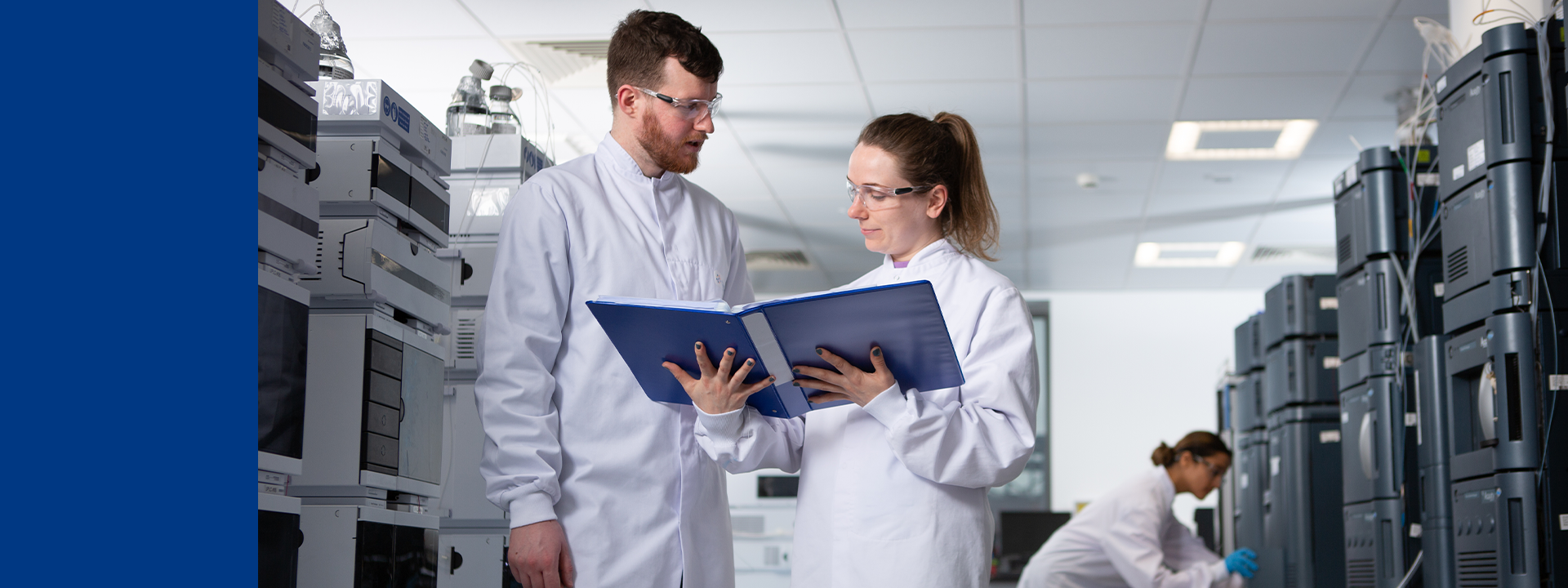 Three individuals in a laboratory setting. Two are standing close together, wearing white lab coats, and looking at a blue folder. The third person, also in a lab coat, is in the background working with laboratory equipment. The lab contains various HLPC&UPLC machines.