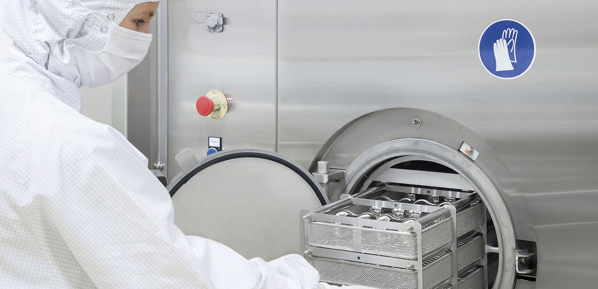 Person in protective clothing operating a large stainless steel industrial machine with an open circular door, revealing trays or containers inside. The machine features a red emergency stop button and a blue safety sign with a hand symbol.