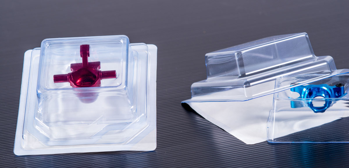 Two transparent plastic packages placed on a dark surface; the left package contains a red object, and the right package contains a blue object.