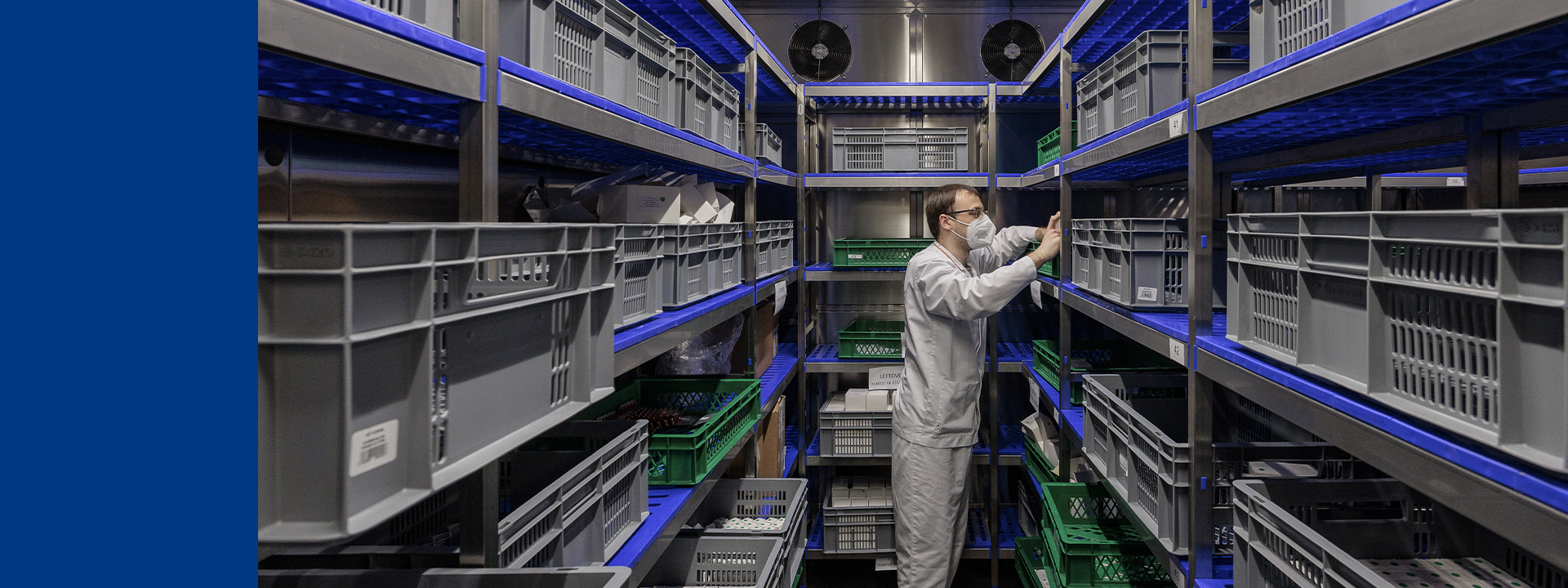 A person in a white protective suit is standing in a stability storage room filled with gray and green plastic crates on metal shelves. The room is illuminated by blue lights, and there are two ventilation fans on the back wall. The person appears to be organising or inspecting the contents of the crates.