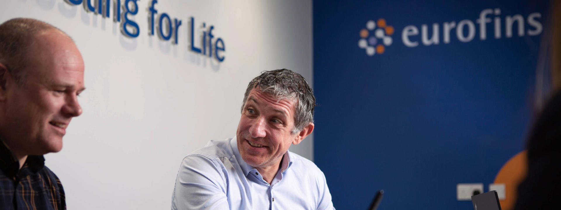 Two consultants engaged in a conversation in a modern office setting, with the Eurofins logo visible in the background. The image captures a collaborative business environment focused on medical devices