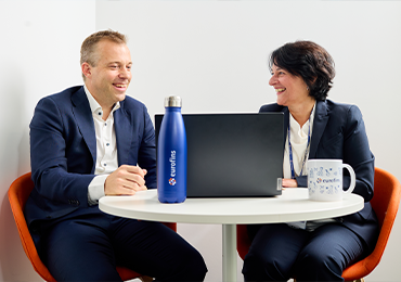 Two people in business attire are sitting at a round table with a laptop, a blue water bottle labeled 'eurofins,' and a white mug also labeled 'eurofins.'