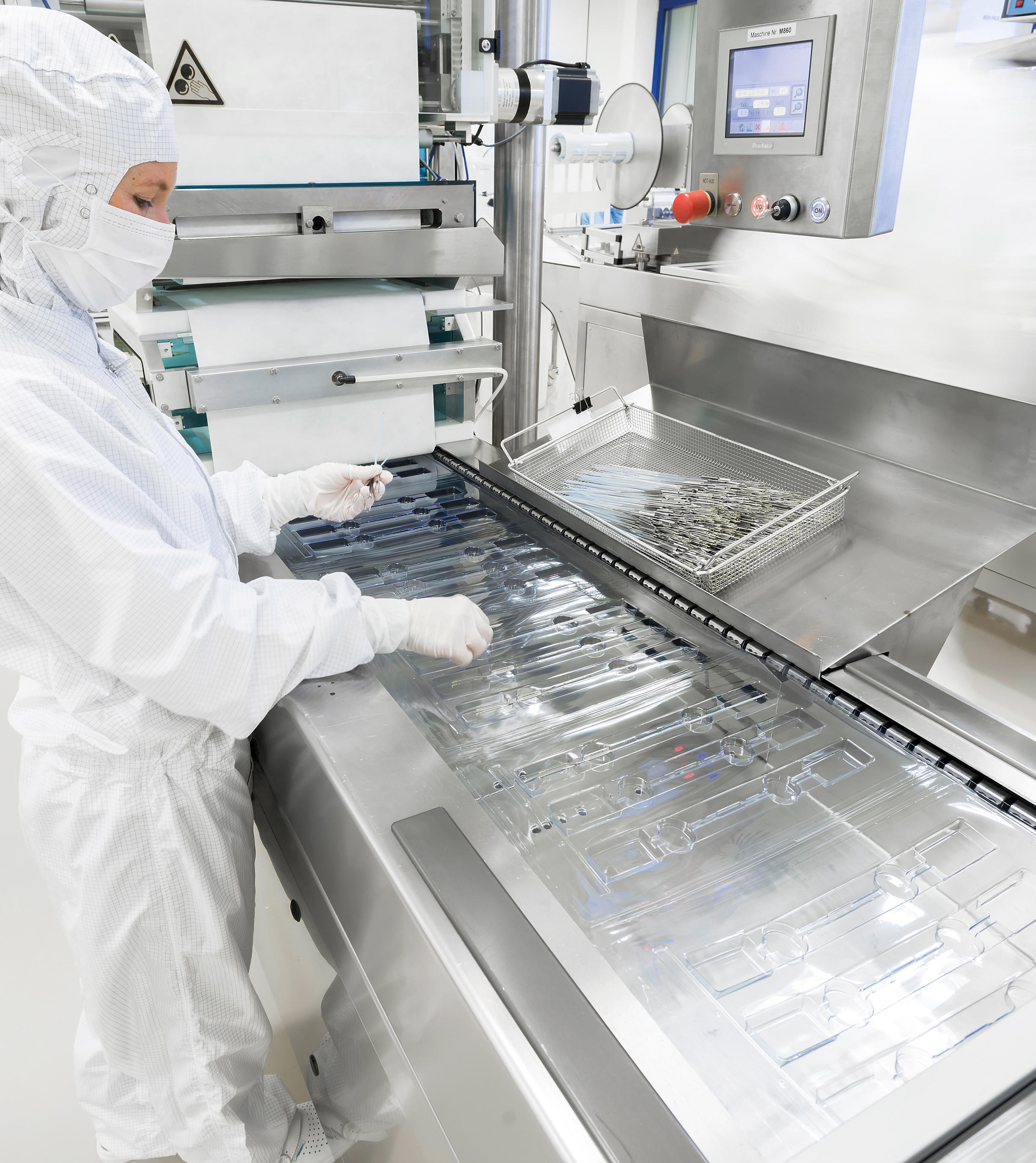Individual in protective clothing working in a cleanroom environment, handling transparent plastic packaging on a conveyor belt amid manufacturing equipment and control panels.