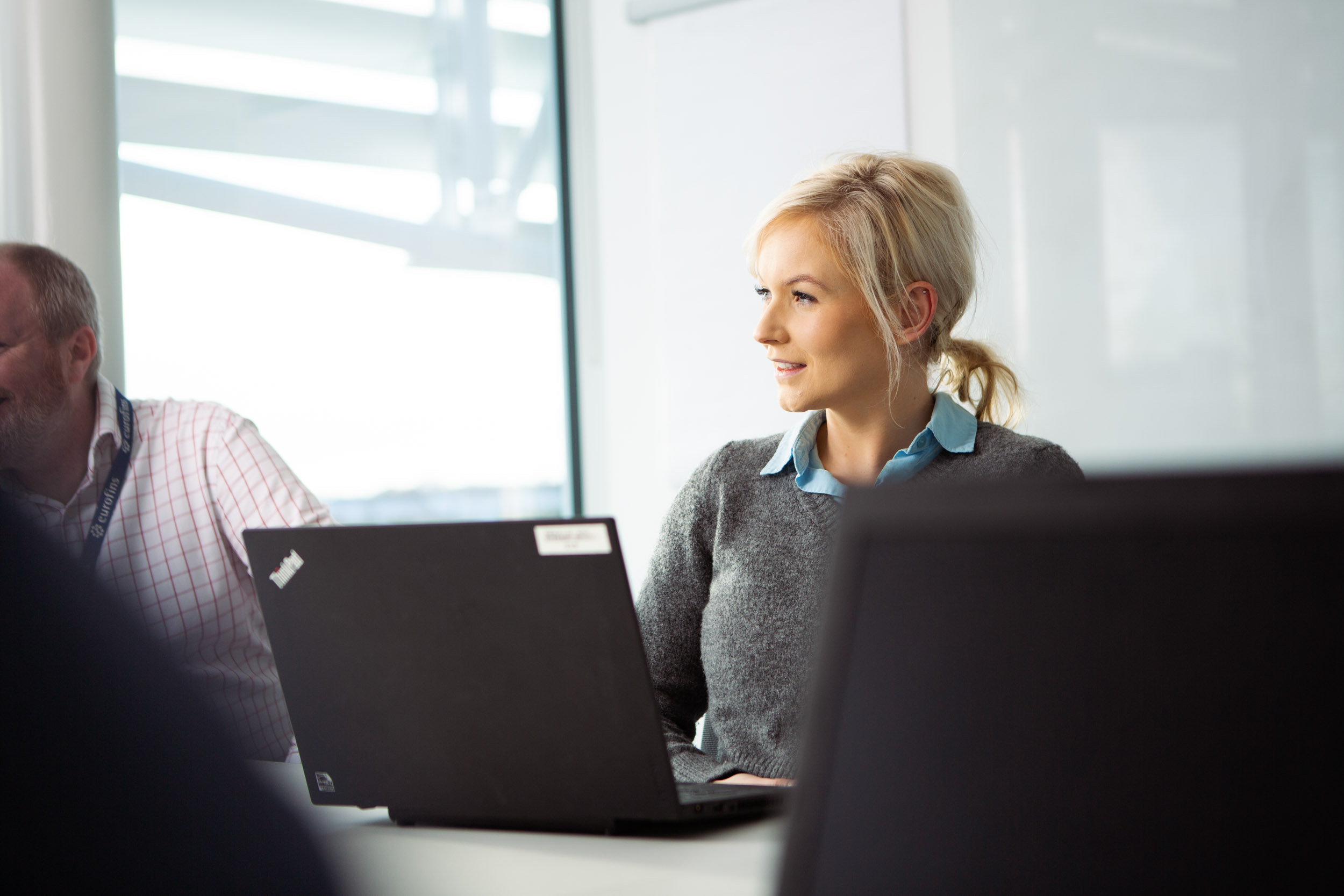 Two people sitting at a table in an office environment, both using laptops. The person on the right wears a grey sweater over a blue collared shirt. Large windows in the background let in natural light