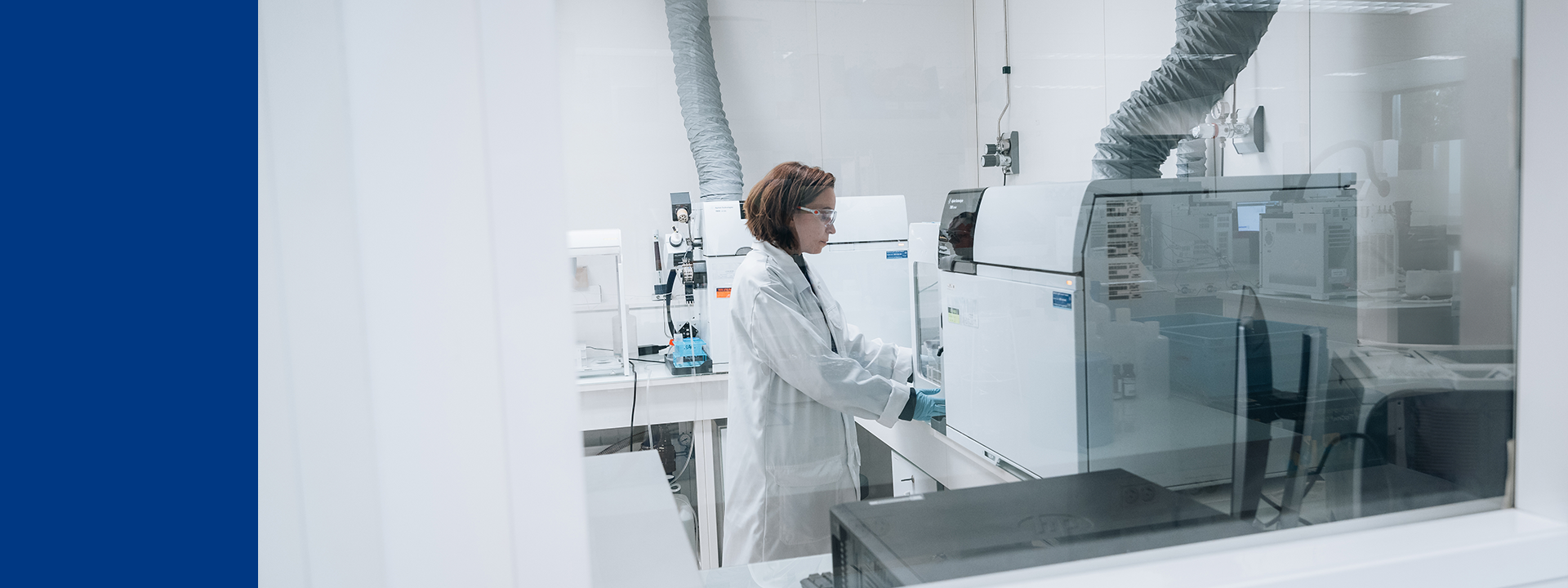 Eurofins BioPharma product Testing laboratory setting with a technician wearing protective clothing, working with scientific equipment. The laboratory has white walls and multiple pieces of equipment, including what appears to be an analytical instrument connected to ventilation ducts. This environment is focused on testing for elemental impurities and trace metals, where advanced analytical techniques are used to detect and quantify trace amounts of metals in pharmaceutical products.