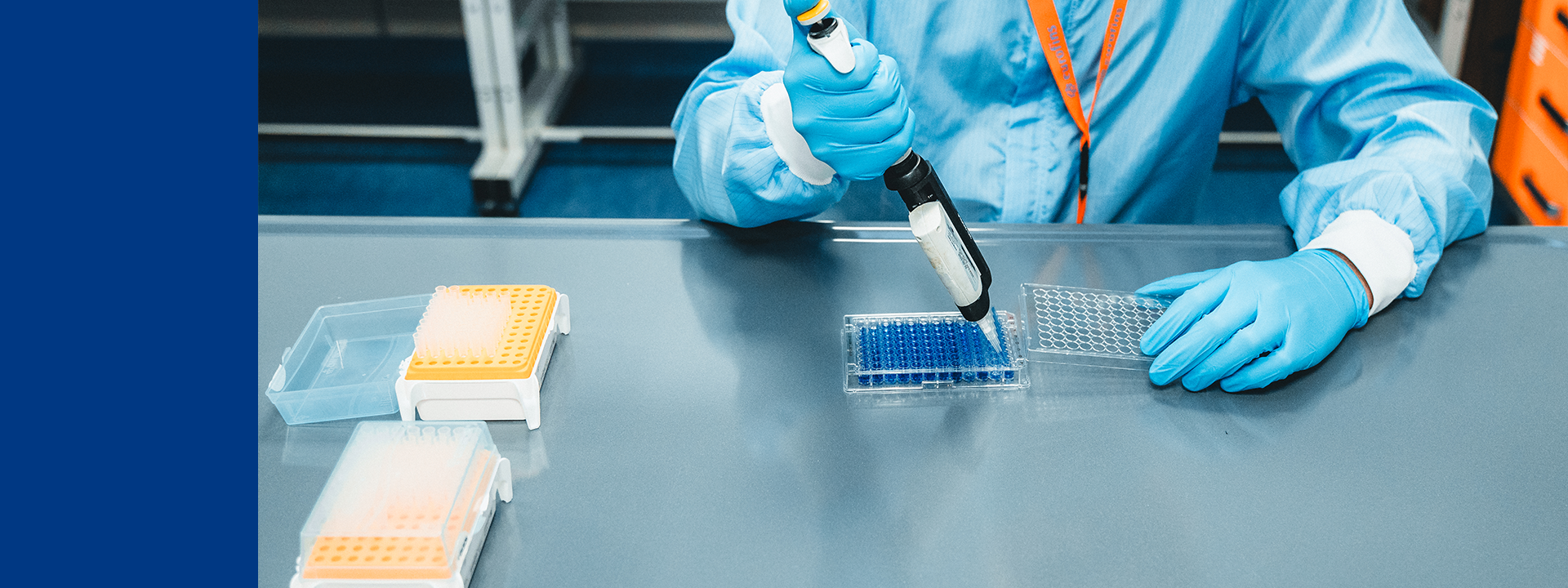 A person wearing blue protective clothing and gloves is using a pipette to dispense liquid into a well plate with multiple wells filled with blue liquid. Two additional well plates with yellow caps are on the table, one partially covered by a transparent lid. Shelving units and laboratory equipment are visible in the background.