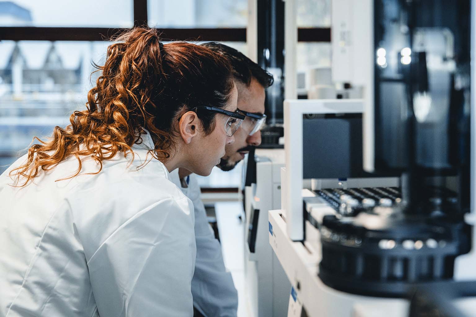 Two individuals in lab coats working together in a laboratory, examining a piece of equipment. The background includes windows with an outside view and various lab instruments
