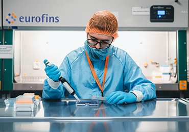 Laboratory setting with a technician wearing protective clothing and gloves, using a pipette to transfer liquid into a container. The background features the 'eurofins' logo and various laboratory equipment. This environment is focused on analytical chemistry and the study of physicochemical properties, which involves analyzing the chemical composition and physical characteristics of substances to ensure their quality, stability, and efficacy in pharmaceutical development.