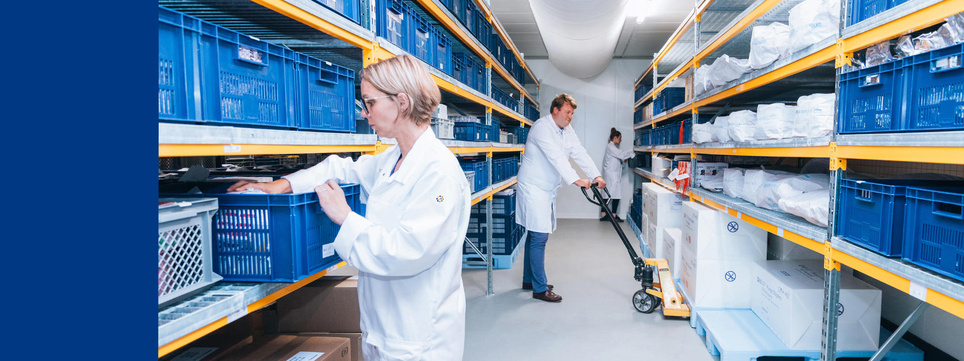 A storage room with shelves filled with blue plastic crates and various items. Two people in white lab coats are organising the crates, while a third person in a white lab coat is using a pallet jack to move boxes. The room is well-lit with overhead lighting and has a clean, organised appearance.