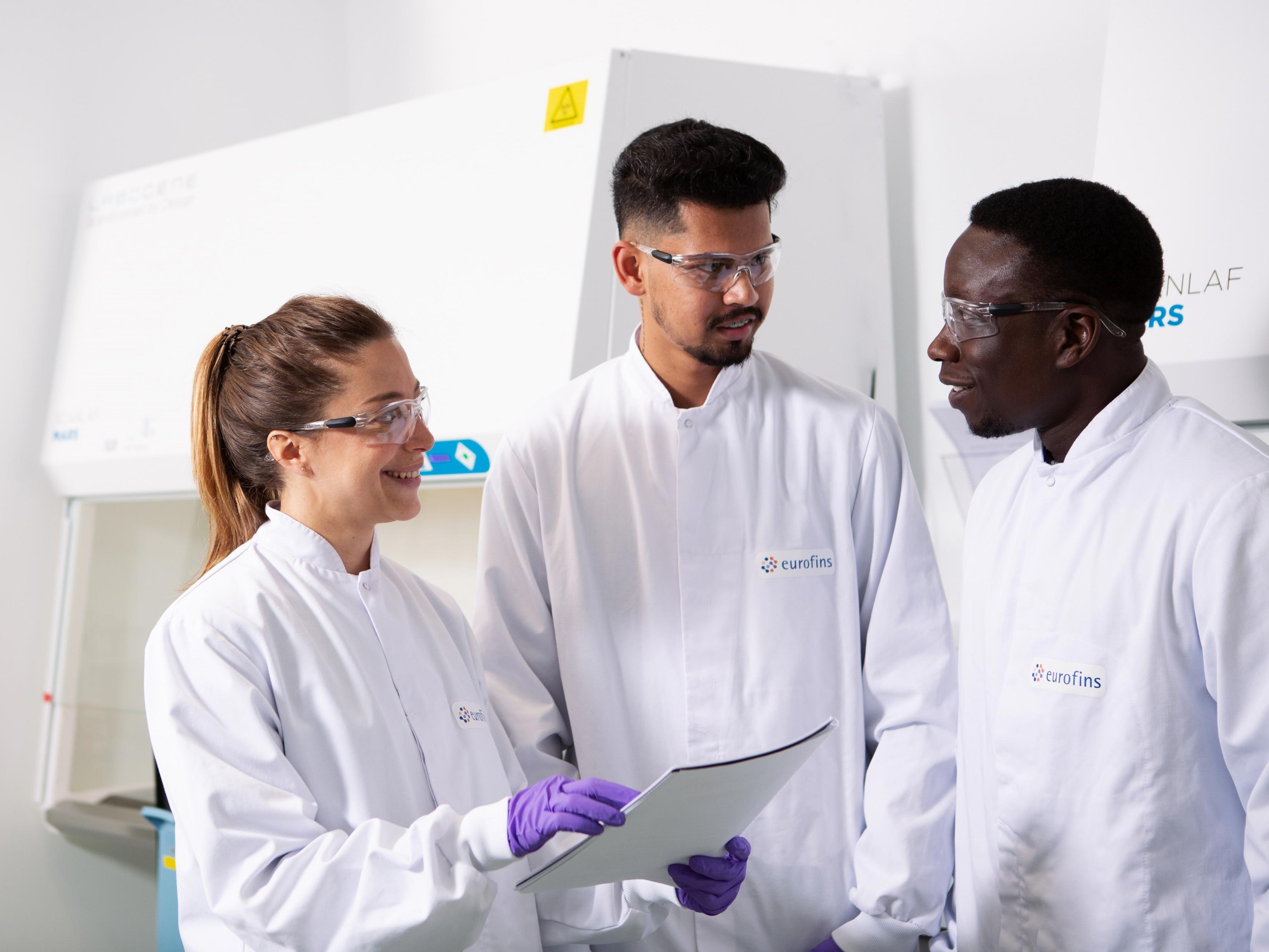 Three individuals wearing white lab coats with the Eurofins logo are standing in a laboratory setting. One person is holding a clipboard, and they appear to be discussing a topic. The background includes laboratory equipment.