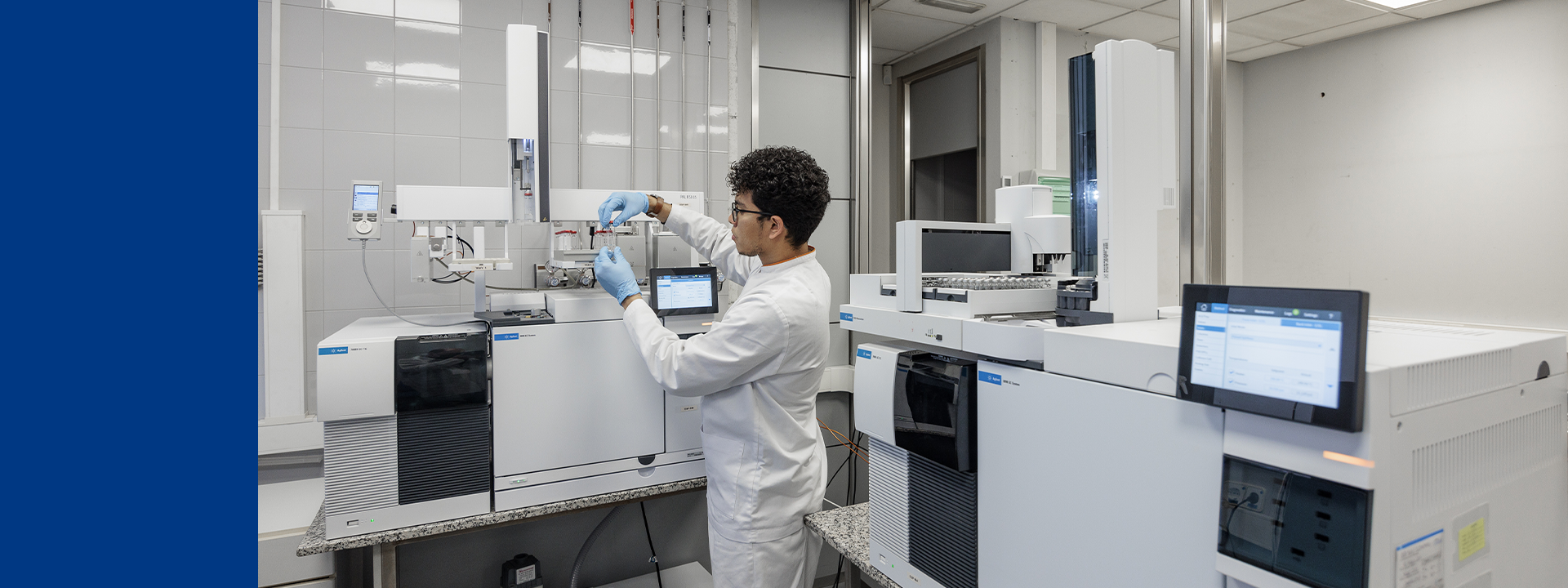 A person in a white lab coat and blue gloves is working with gas chromatograph equipment in a laboratory. The person is adjusting or placing something into one of the machines, which are large, white, and equipped with various screens and compartments. The laboratory has a clean, organised appearance with tiled walls and ceiling lights.