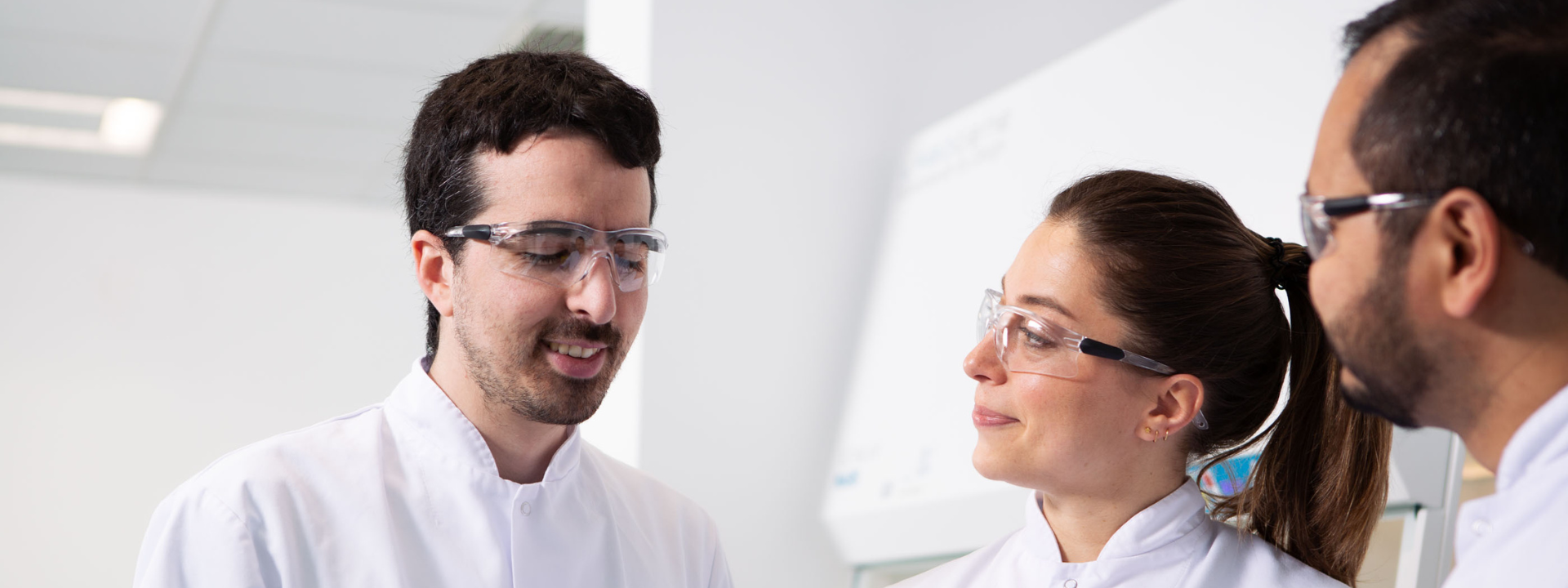 A group of three people wearing white lab coats are standing and talking in a laboratory setting. Their faces are blurred