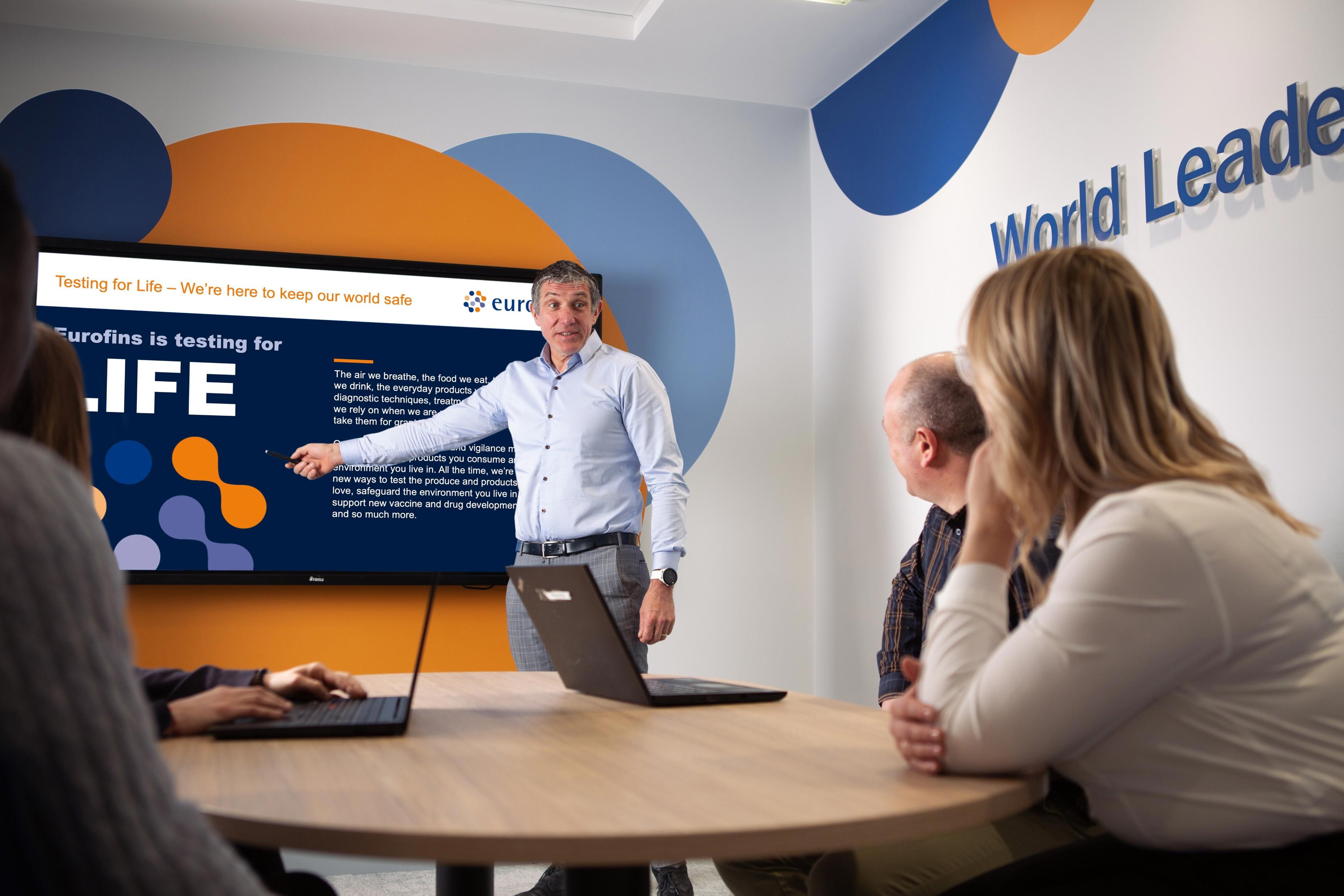 A man in a blue shirt is standing and pointing at a large screen during a presentation in a modern conference room. The screen displays the title "Eurofins is testing for LIFE" and some text about testing for life to keep the world safe. Four people are seated around a table with laptops, attentively listening to the presenter. 