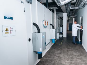 A person in a white lab coat is operating equipment in a storage ATEX with several large, gray machines mounted on the wall. The machines have various pipes and cables connected to them. There are warning signs and labels on the walls and machines.