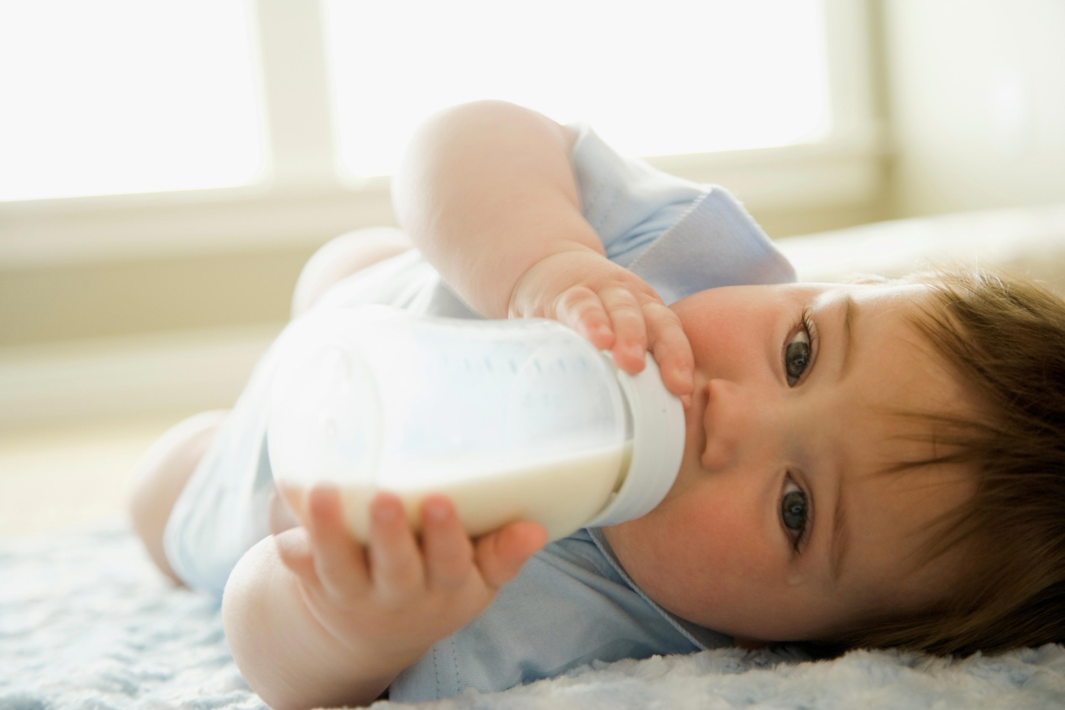 A baby is lying on a soft, light-coloured surface, holding a bottle filled with milk. The baby is dressed in light-coloured clothing and appears to be in a relaxed position. The background shows a bright room with large windows allowing natural light to fill the space.