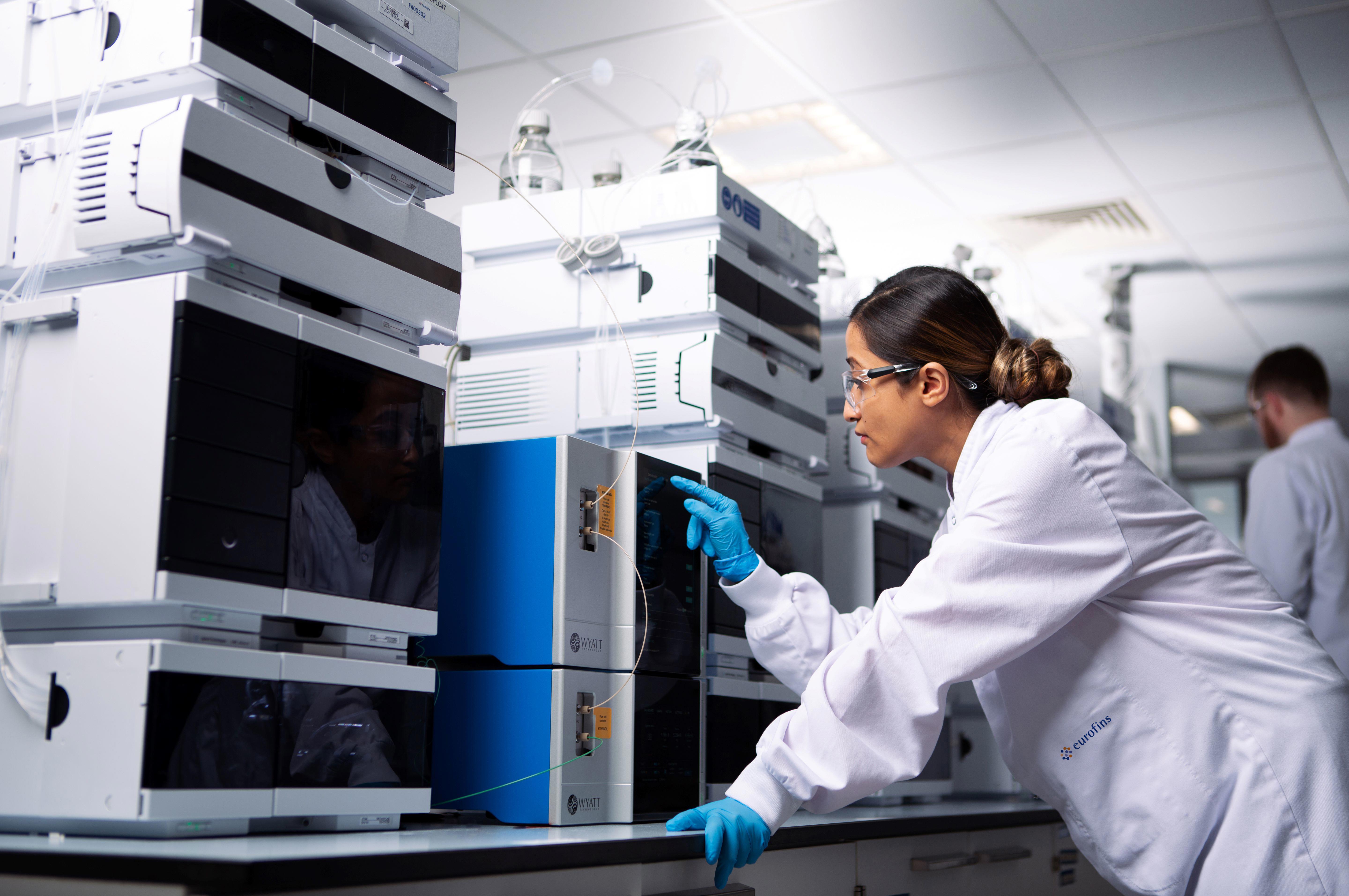 A laboratory technician wearing a white lab coat and blue gloves operates an ICE imaged capillary electrophoresis equipment in a modern laboratory. The technician is adjusting settings on the equipment, surrounded by other similar machines with various cables and tubes connected. Another person in the background is also working in the lab.
