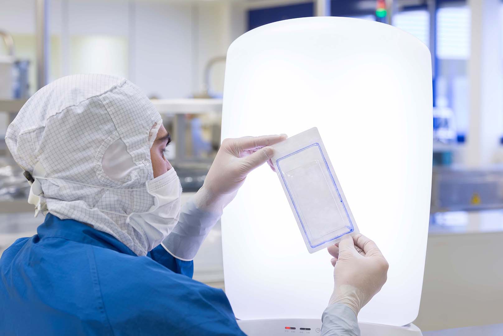 Person in a cleanroom suit, including a hair cover and face mask, holding a sealed package up to a bright light source in a laboratory or cleanroom environment with equipment and workstations in the background.