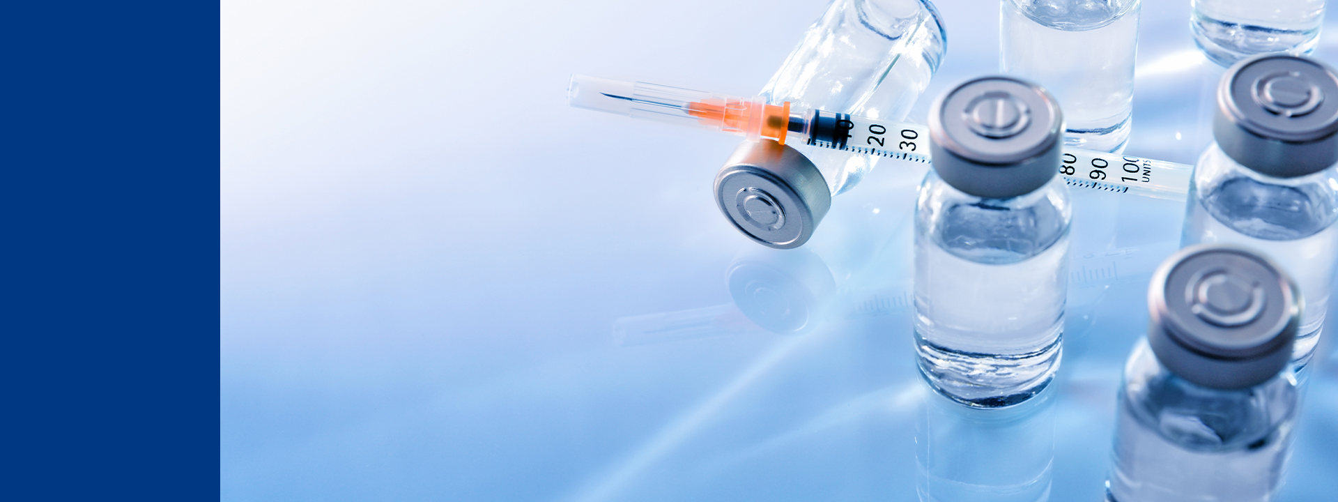 A syringe marked with measurement units rests across a sealed vial filled with clear liquid, surrounded by similar vials with gray caps. Set against a clean, light blue background, the image reflects the precision and sterility required for syringe testing in regulatory and market readiness evaluations.