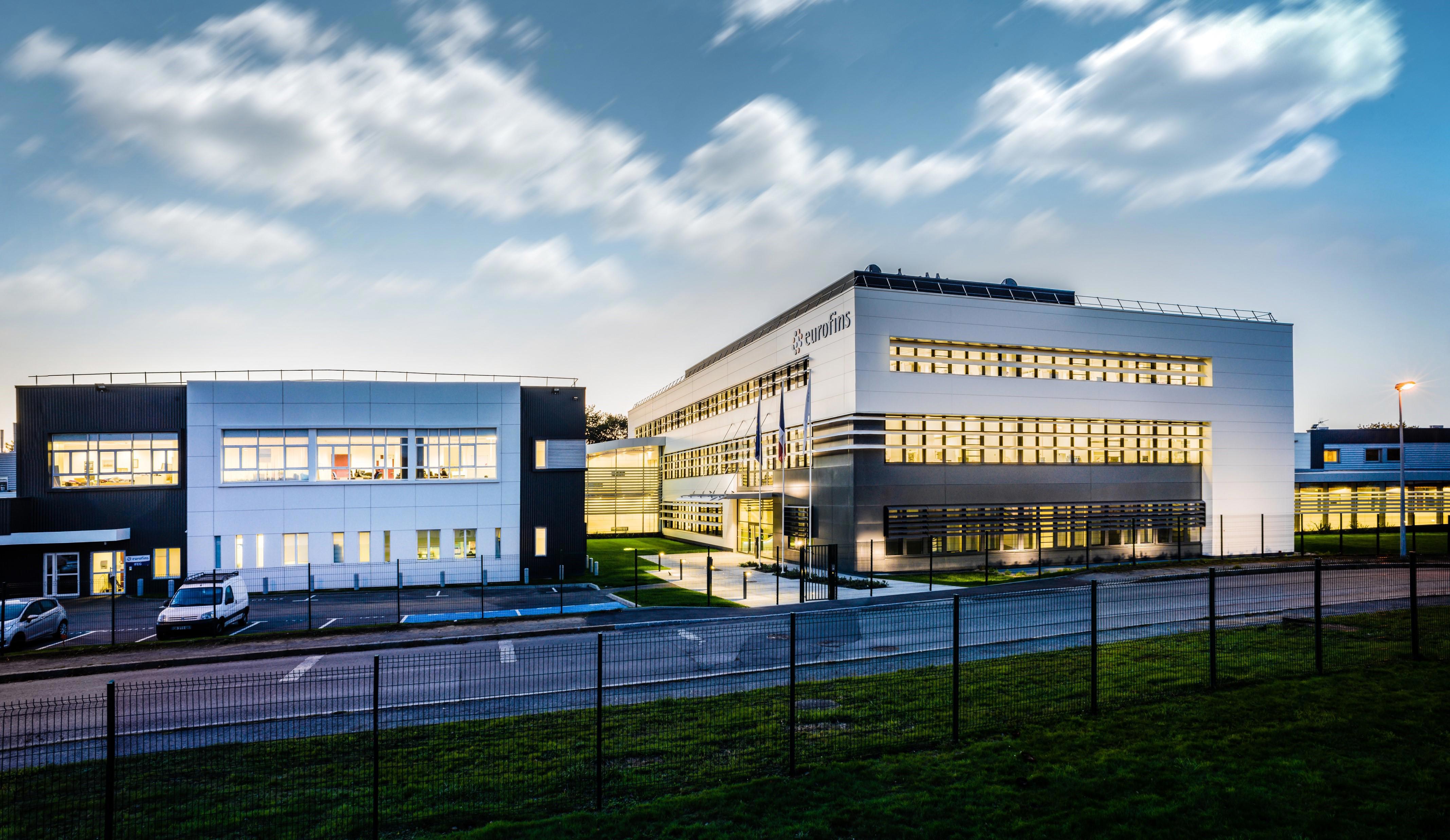 The Eurofins laboratory in Nantes, France, featuring two modern buildings with large windows, illuminated during twilight. The buildings are surrounded by a well-maintained lawn and a fence in the foreground. The sky is partly cloudy, and several cars are parked near the buildings.