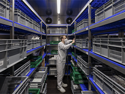 A person in a white protective suit is standing in a storage room filled with gray and green plastic crates on metal shelves. The room is illuminated by blue lights, and there are two ventilation fans on the back wall. The person appears to be organising or inspecting the contents of the crates.