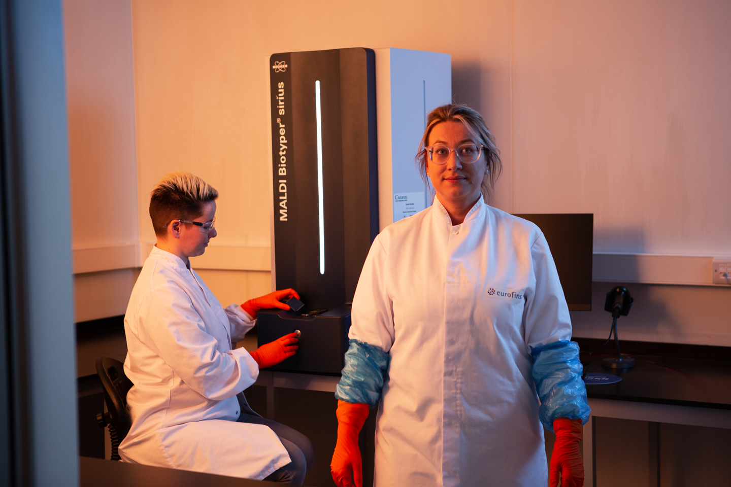 Two scientists in a lab working with a MALDI-TOF instrument, illustrating advanced technology used in the production and banking of microbial strains.