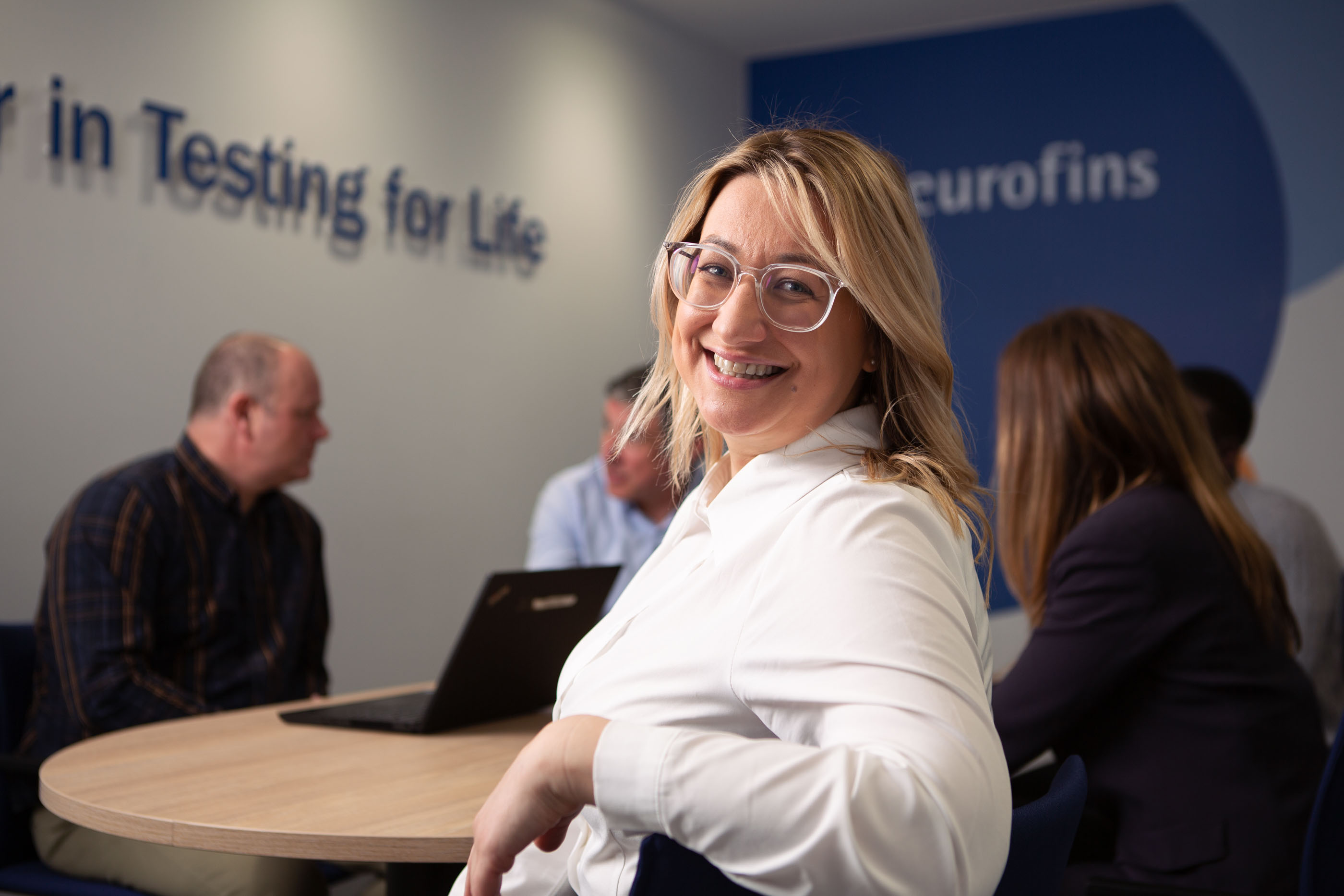 Group of people sitting around a table in an office or meeting room with 'Eurofins' and 'Testing for Life' text on the wall. Four individuals are visible with blurred faces; one in the foreground wears a white shirt, and others are using laptops or conversing