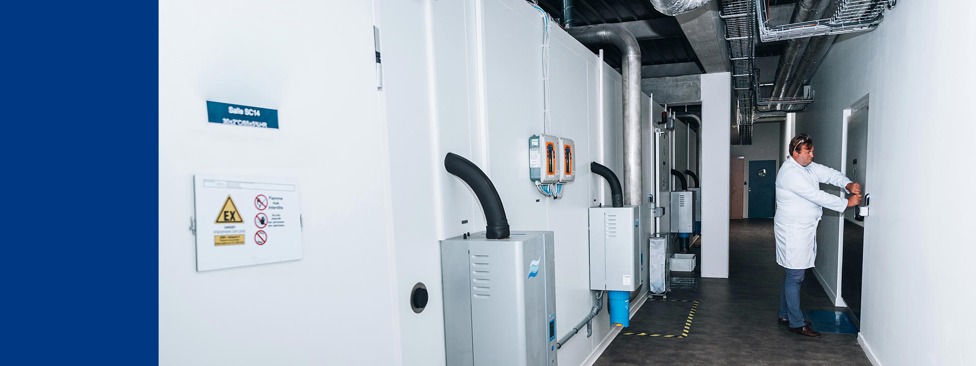 A person in a white lab coat is operating equipment in a storage ATEX with several large, gray machines mounted on the wall. The machines have various pipes and cables connected to them. There are warning signs and labels on the walls and machines.