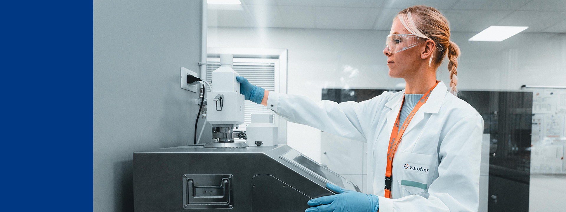 Eurofins BioPharma Product Testing laboratory setting with a technician wearing a white lab coat and blue gloves, operating or testing equipment. The technician is holding a device connected to the equipment on the counter. This environment is focused on container testing, which involves evaluating the physical and chemical properties of containers to ensure they meet the required standards for protecting and preserving pharmaceutical products.