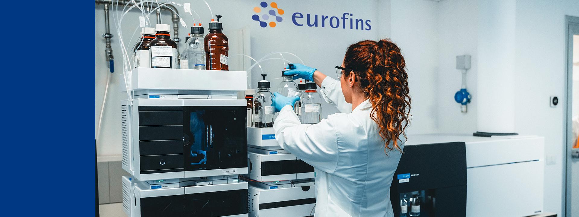 A scientist in a white lab coat and blue gloves operates a complex analytical instrument connected to chemical bottles in a Eurofins laboratory. The equipment, featuring stacked modules with digital displays and tubing, is used for toxicological risk assessment in Extractables & Leachables testing.