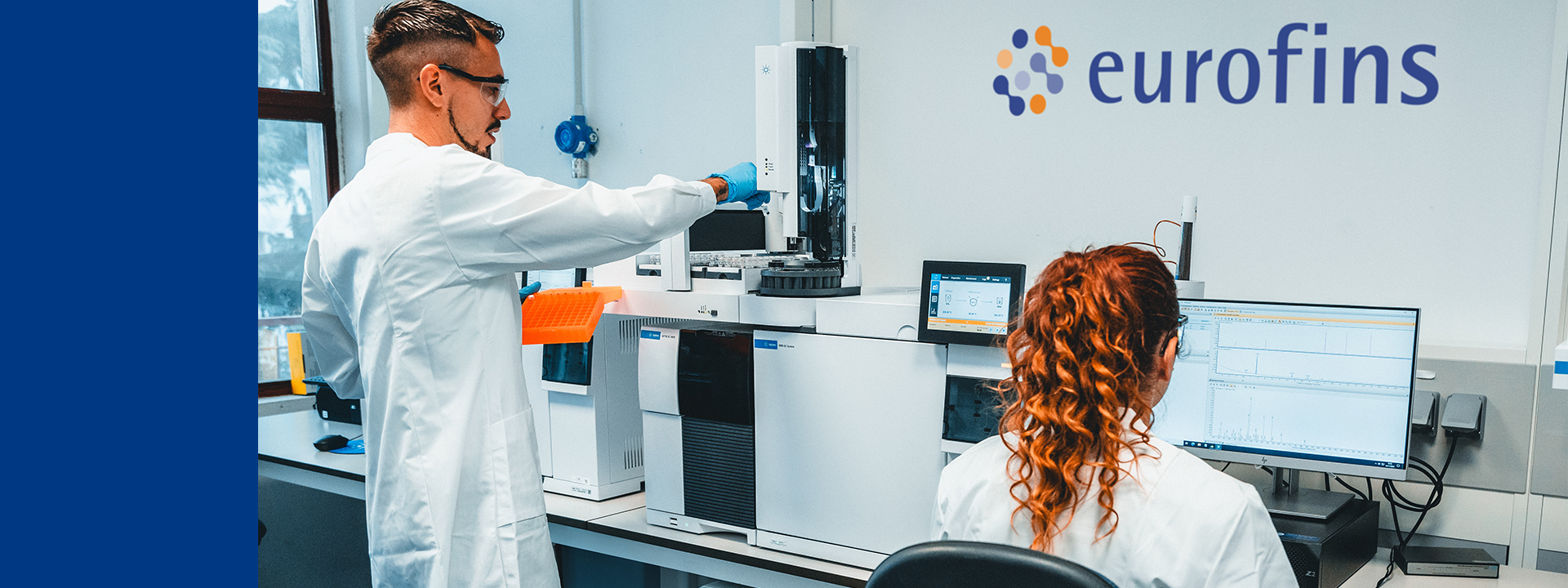 Two analysts in a Eurofins BioPharma Product Testing laboratory setting. One individual is standing and operating a piece of laboratory equipment, while the other is seated at a computer workstation. The laboratory contains various pieces of equipment and instruments, including what appears to be a chromatograph or similar analytical device. This work is part of the comparator product testing conducted at Eurofins BioPharma Product Testing.