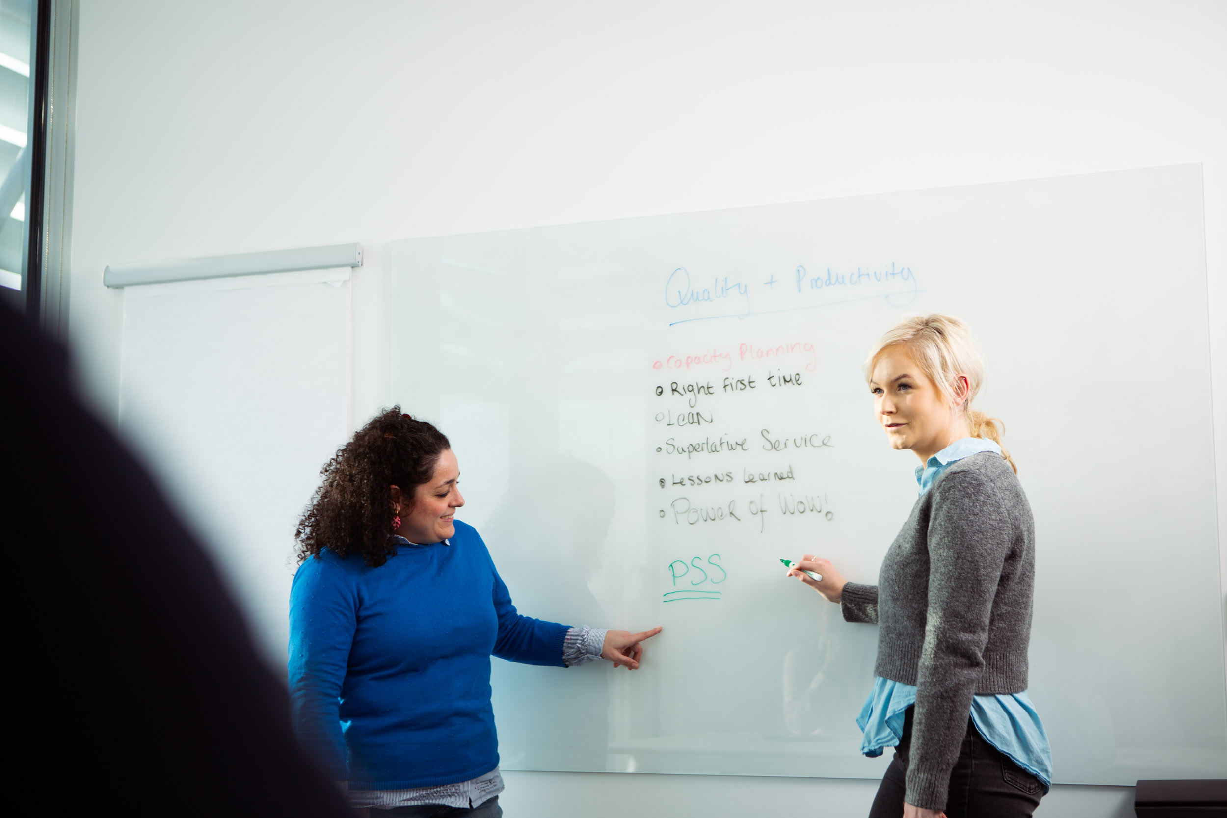 Two individuals standing in front of a whiteboard with handwritten text including 'Quality + Productivity,' bullet points such as 'Project Planning,' 'Right first time,' 'Lean,' 'Supportive Service,' 'Lessons learned,' 'Proud of work,' and the acronym 'PSS