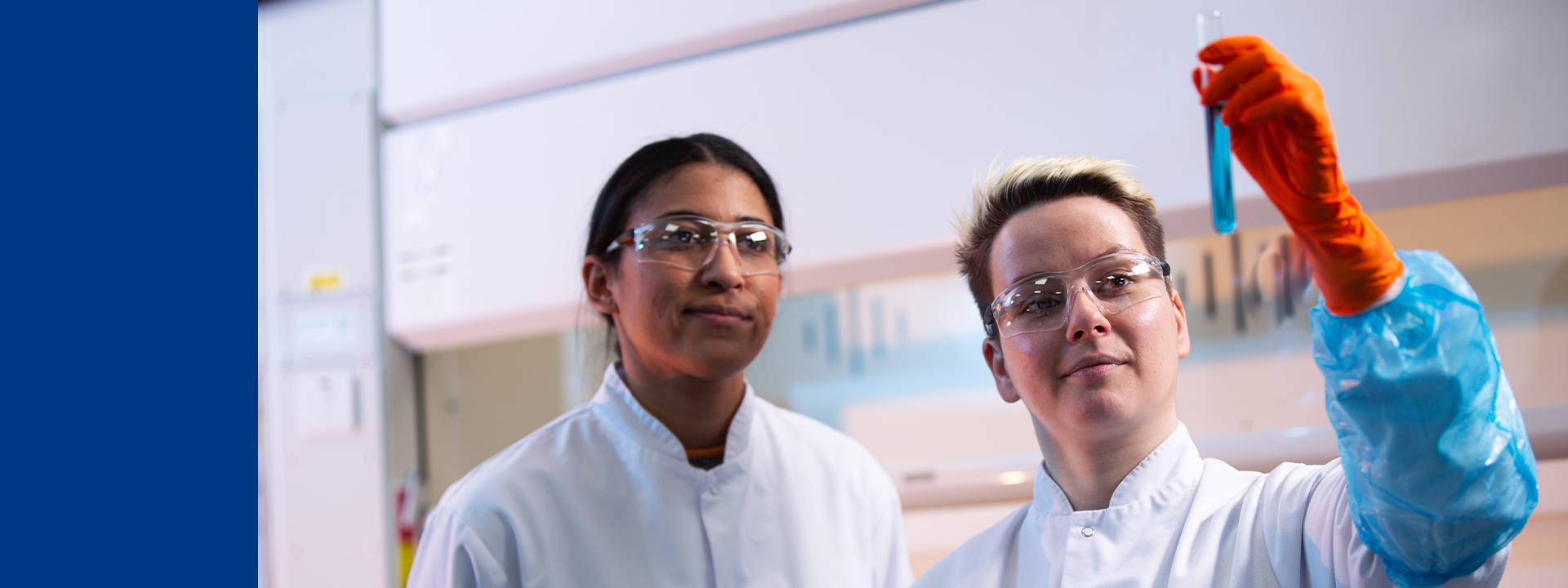 Two laboratory technicians in a laboratory setting, both wearing white lab coats. One technician is holding up a test tube containing a blue liquid, wearing an orange glove on the hand holding the test tube. The background includes laboratory equipment and cabinets, indicating an environment focused on analytical development and product characterization at Eurofins BioPharma Product Testing.