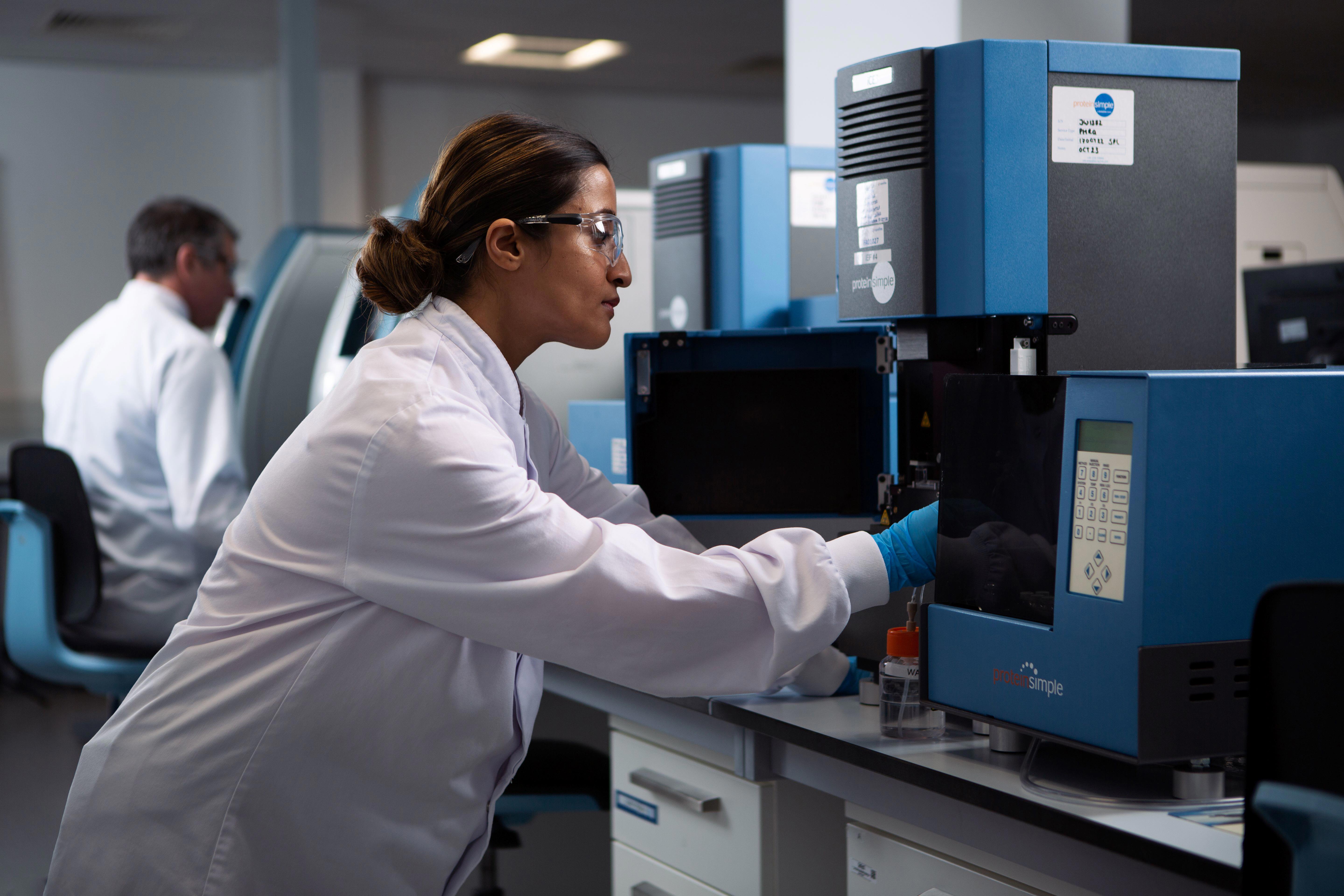 A laboratory technician wearing a white lab coat and blue gloves is operating a blue and black ICE imaged capillary electrophoresis instrument on a countertop. Another person in the background is also working with similar equipment.