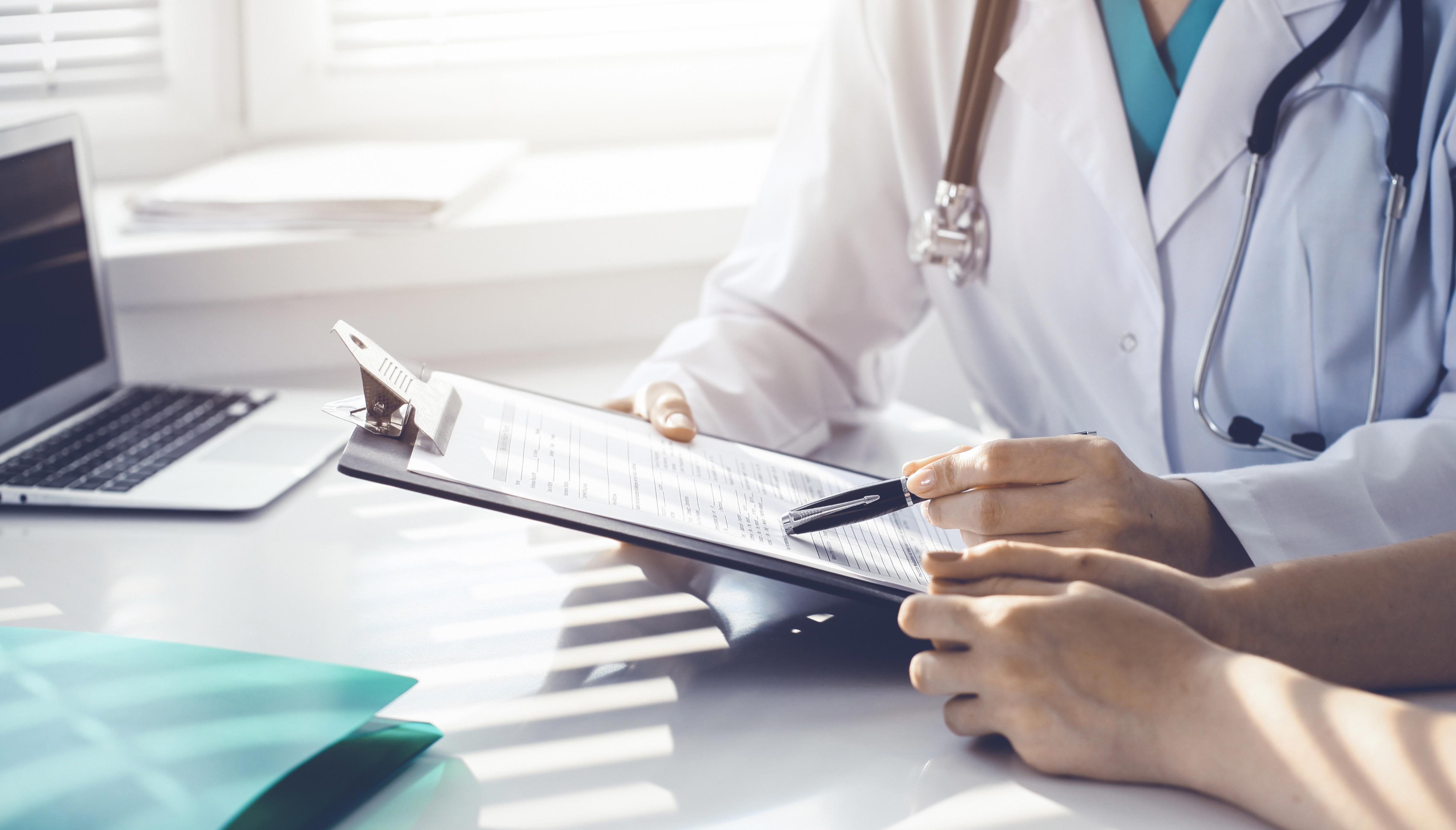 A doctor in a white coat with a stethoscope is holding a clipboard and pointing at it with a pen. Another person, whose hands are visible, is sitting across the desk. There is an open laptop and some documents on the desk.
