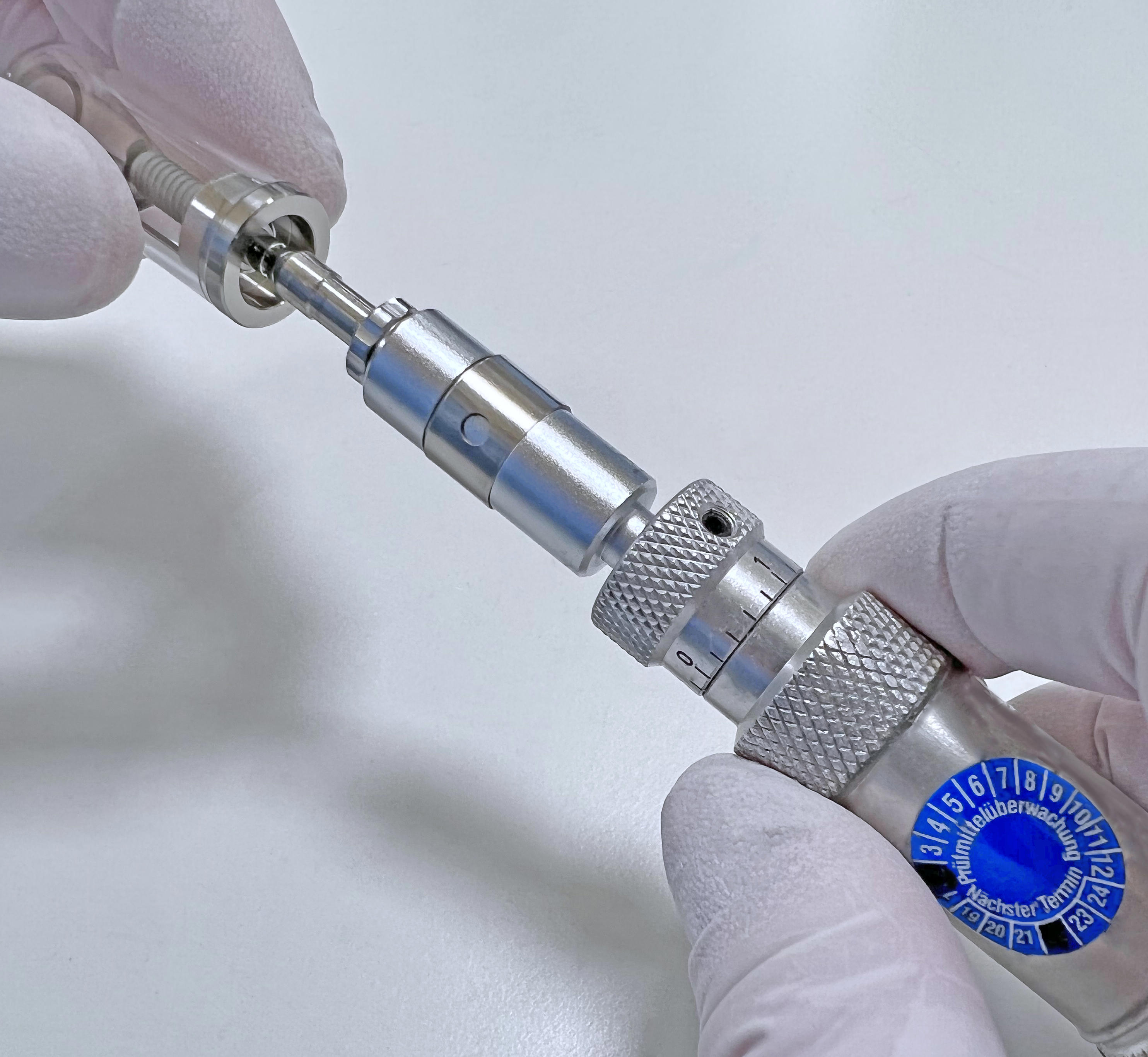 Close-up of a person wearing white gloves holding a metallic precision tool with both hands. A blue circular dial with numbers from 0 to 12 is visible on the right side, likely part of the tool's adjustment mechanism