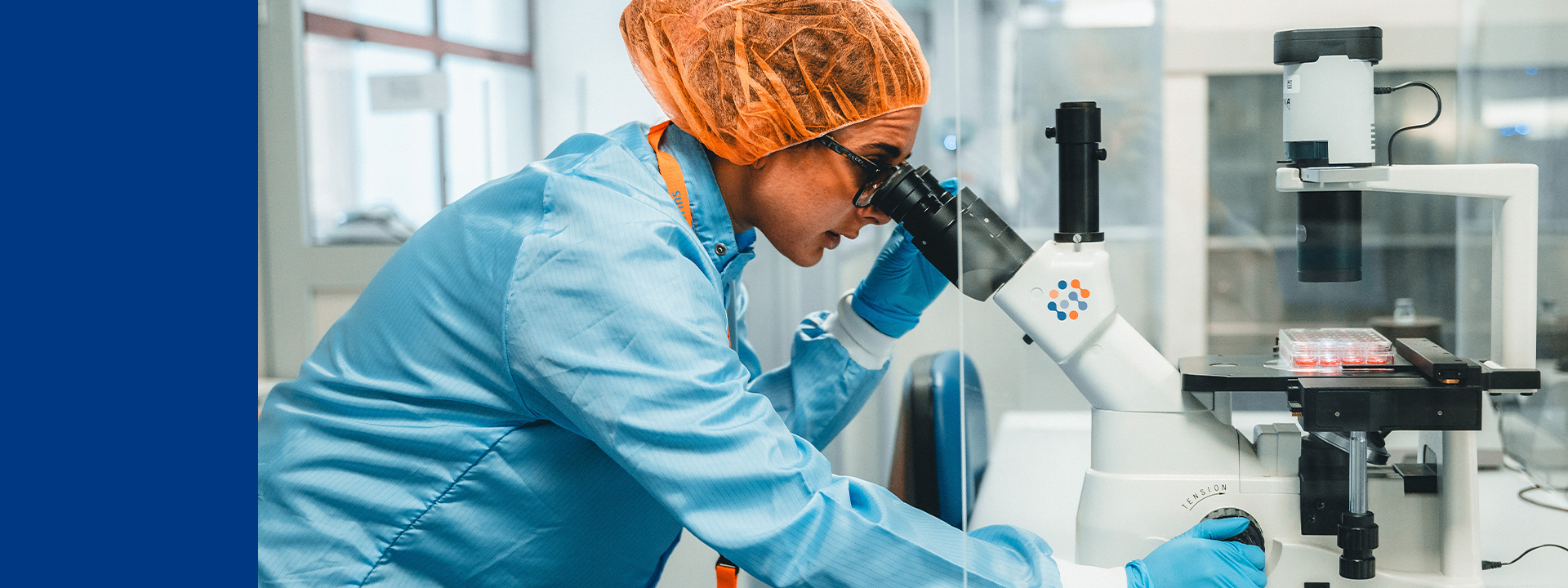 A laboratory technician in a blue lab coat, orange hairnet, and blue gloves examines samples under a microscope branded with the Eurofins logo. The setting highlights analytical procedures used in the characterisation of large molecules.