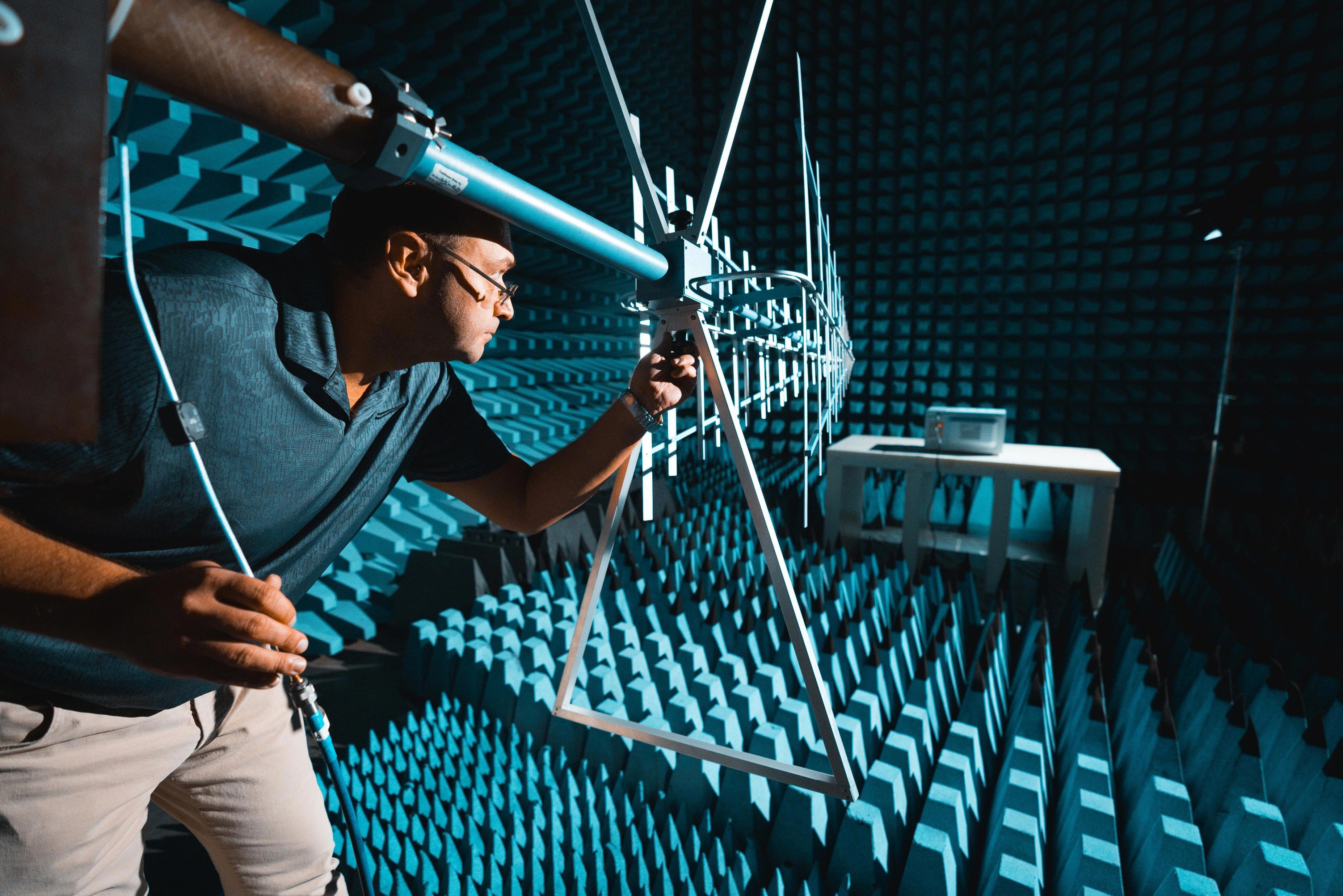 A person is inside an anechoic chamber, which is a room designed to completely absorb reflections of sound or electromagnetic waves. The walls, ceiling, and floor are covered with blue foam pyramids that help in absorbing sound waves. The person is holding a large antenna-like structure and appears to be conducting some sort of experiment or test.