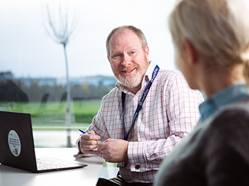 A man wearing a checkered shirt and a eurofins ID lanyard, is sitting at a table with a laptop in front of him. He is holding a pen and appears to be engaged in conversation with another person whose back is facing the camera. The background shows an outdoor scene with greenery and trees.
