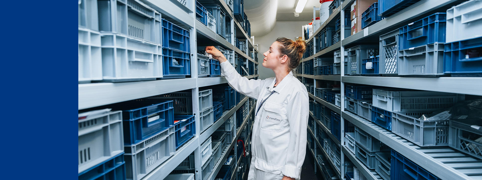 A person wearing a white lab coat with the logo 'eurofins' is standing in an aisle between shelves filled with blue and gray plastic storage crates. The person is reaching into one of the crates on the left side.