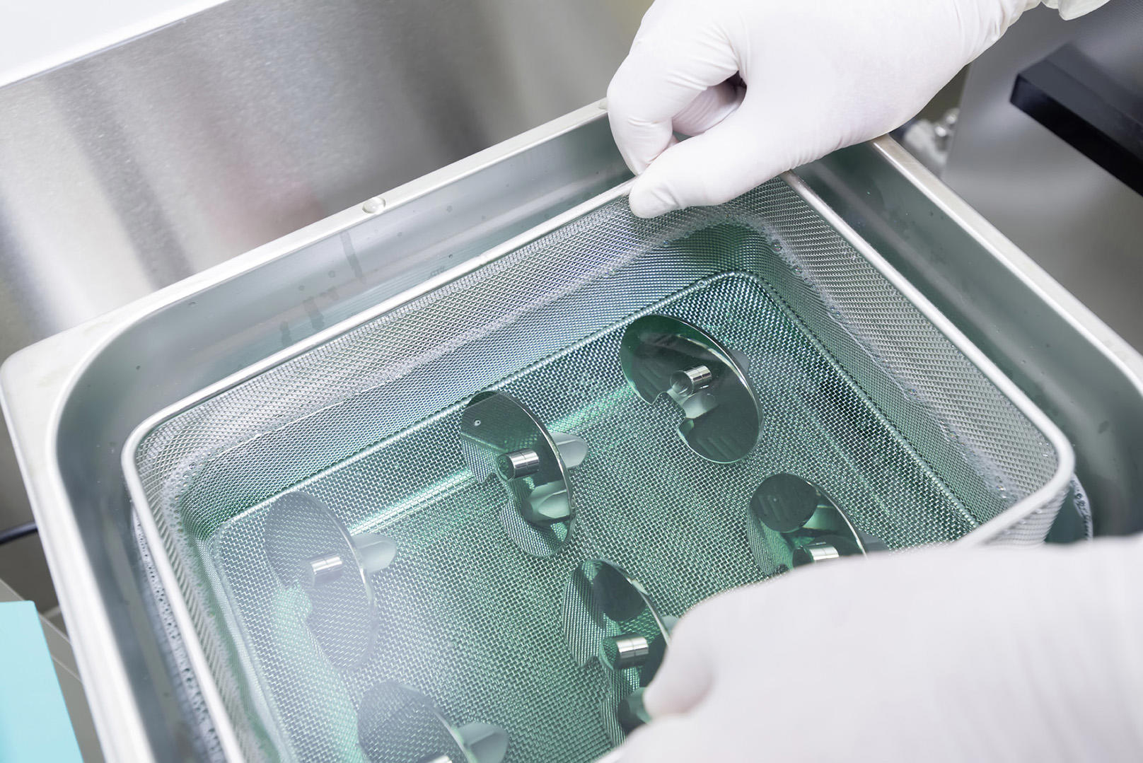 A pair of gloved hands handling a metal mesh basket containing several small, reflective devices submerged in a liquid within a stainless steel container. The image is labeled 'Active Devices'