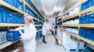 Stability Storage room with shelves filled with blue plastic baskets. Three people in white lab coats are working: one handling a basket, another using a pallet jack, and a third person further back. The shelves are organised with various items stored in the baskets.