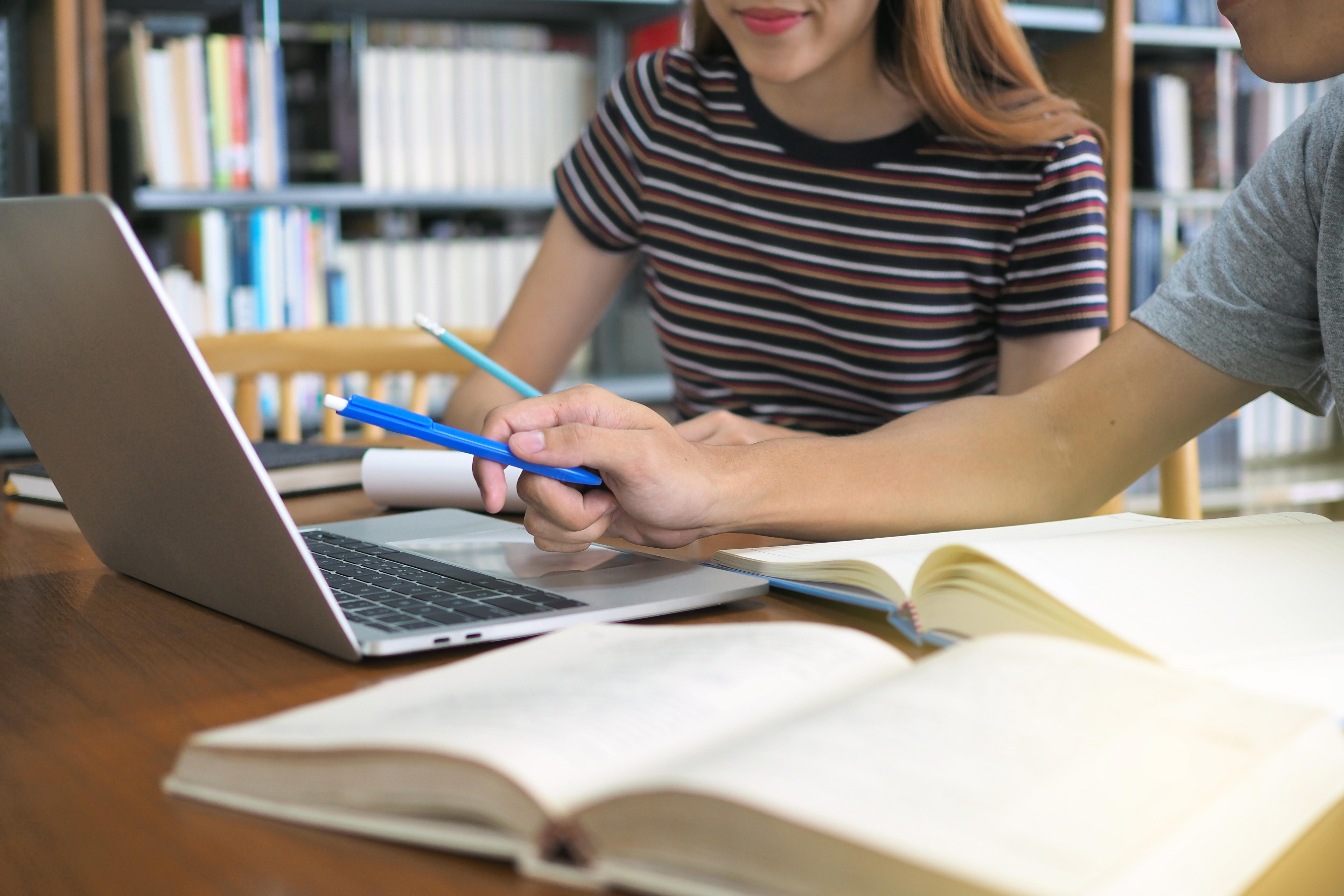 Two people are sitting at a wooden table in a library or study area, working together on a laptop. One person is pointing at the laptop screen with a blue pen while the other person looks on. Open books and notebooks are spread out on the table in front of them.