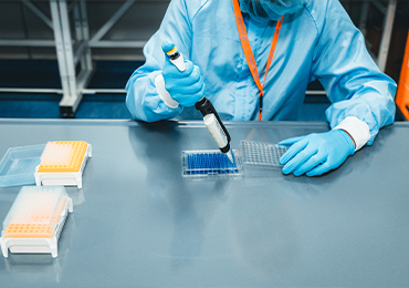 A scientist in a white blue coat and blue gloves operates a complex analytical instrument connected to chemical bottles in a Eurofins laboratory. The equipment, featuring stacked modules with digital displays and tubing, is used for toxicological risk assessment in Extractables & Leachables testing.