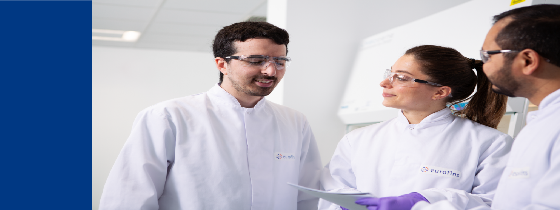 Three laboratory scientists in white lab coats and protective eyewear discussing documents in a microbiology lab, representing collaboration in the production and banking of microbial strains.