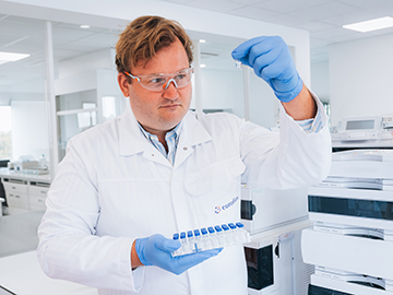 A scientist in a laboratory is holding a blue-capped test tube rack with multiple test tubes. The scientist is wearing a white lab coat with the logo "eurofins", blue gloves, and has short hair. The background shows laboratory equipment and workstations.