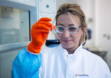 A scientist in a white lab coat bearing the Eurofins logo and wearing orange protective gloves holds up a flask containing a dark liquid for visual inspection. The laboratory background, equipped with scientific instruments and blurred to emphasize the subject, reflects a controlled environment dedicated to analytical method development and validation processes at Eurofins BioPharma Product Testing.
