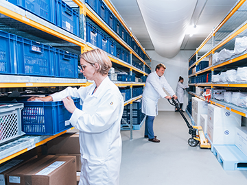 A storage room with shelves filled with blue plastic crates and various items. Two people in white lab coats are organising the crates, while a third person in a white lab coat is using a pallet jack to move boxes. The room is well-lit with overhead lighting and has a clean, organised appearance.