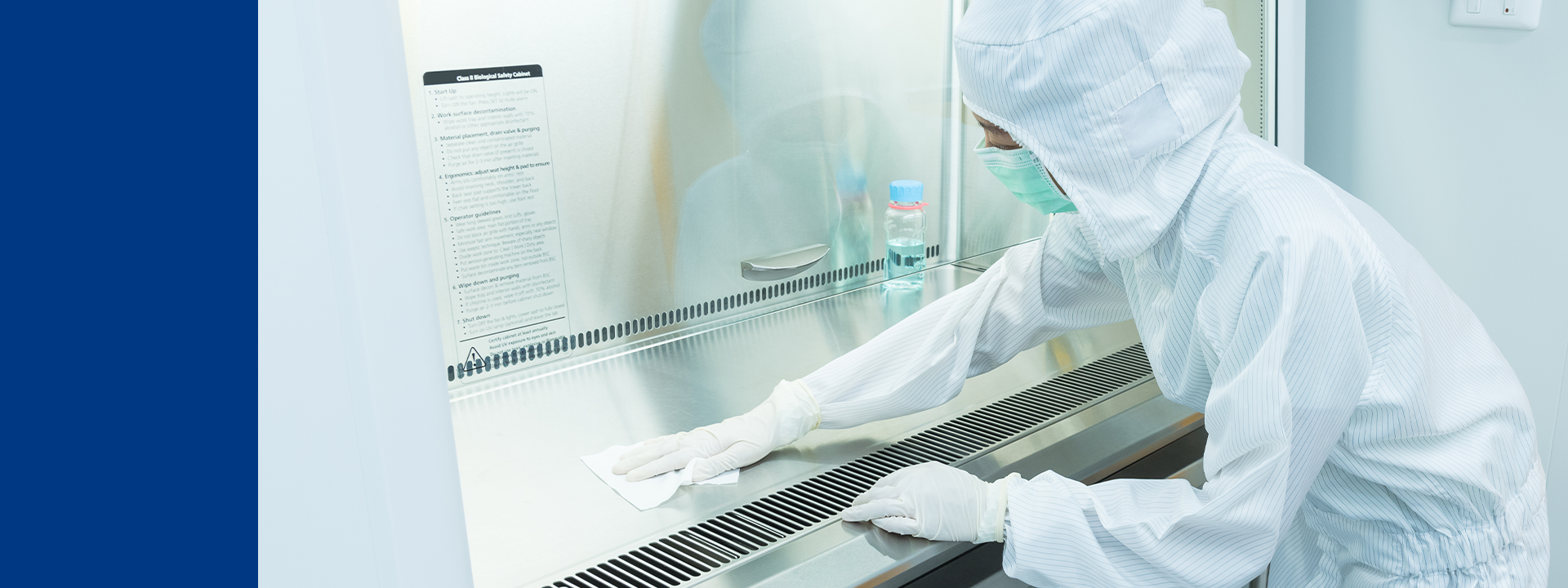 A person wearing full protective gear, including a hooded suit, gloves, and a face mask, is cleaning the surface of a biosafety cabinet. Several bottles and containers are visible inside the workstation.