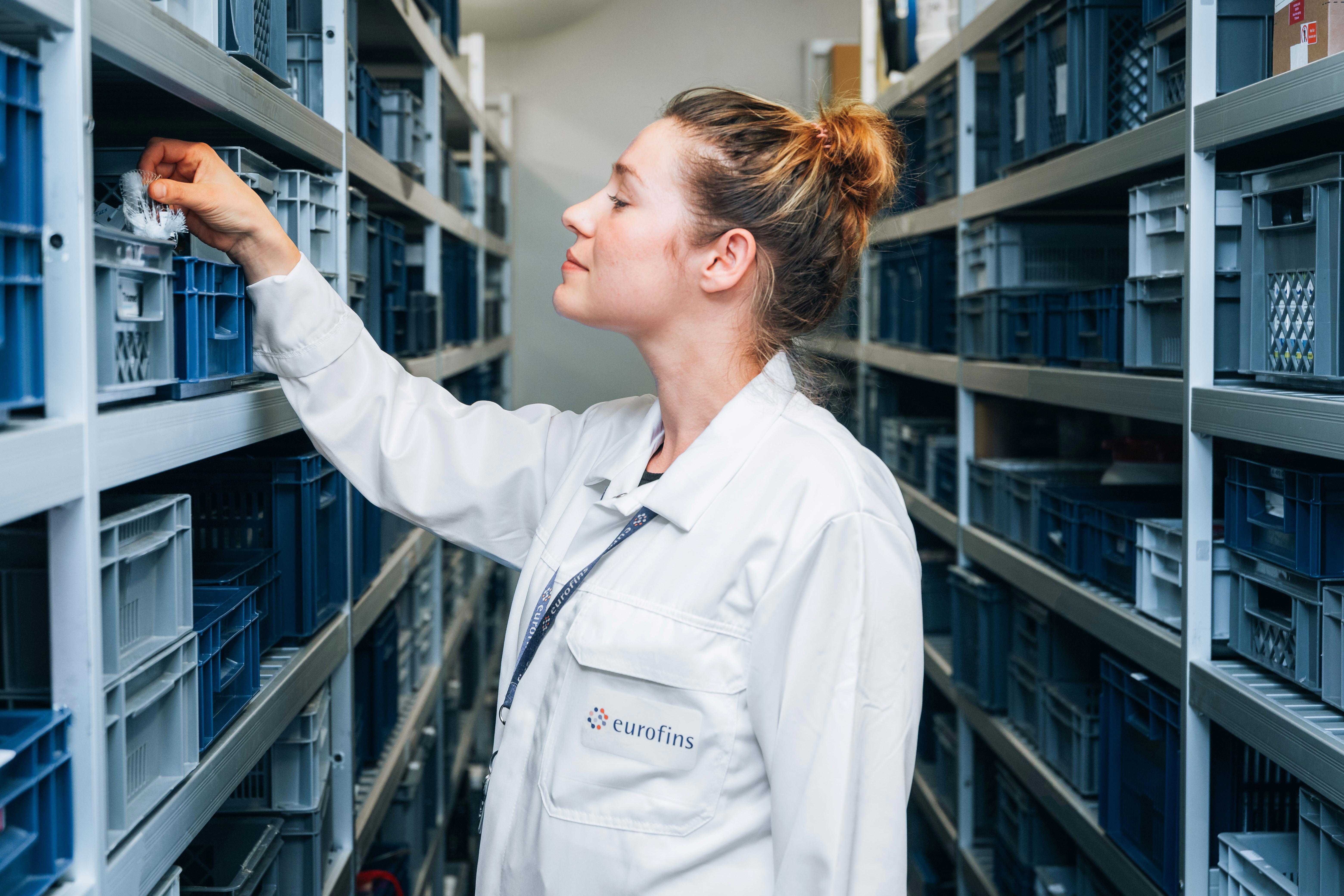 A person wearing a white lab coat with the logo 'eurofins' is standing in an aisle between shelves filled with blue and gray plastic storage crates. The person is reaching into one of the crates on the left side.